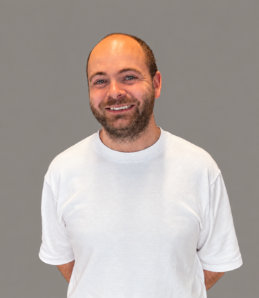 Smilende mann i hvit T-skjorte mot grå bakgrunn, portrettbilde med avslappet positur.