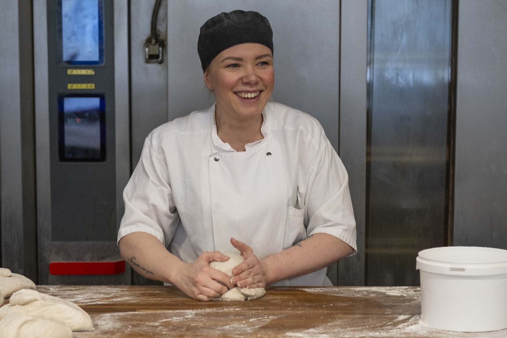 Smilende baker i hvit arbeidsuniform elter deig på kjøkkenet ved en stor bakebenk
